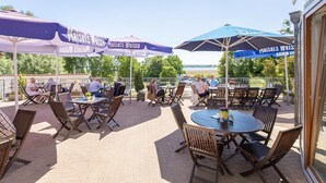 Een terras buiten met tafels en stoelen onder parasols, met uitzicht op een waterpartij.