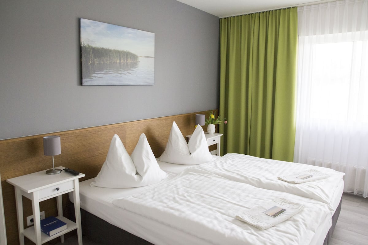 Een hotelkamer met een groot bed, een nachtkastje met een lamp, een klein tafeltje, een schilderij aan de muur en groene gordijnen.