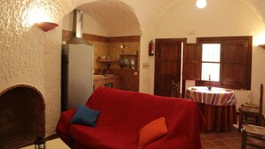 A cozy living room with a red sofa, a fireplace, and a kitchen area with wooden cabinets and a round table. - Las Chumberas Cave - Los Cerezos (Benalua)