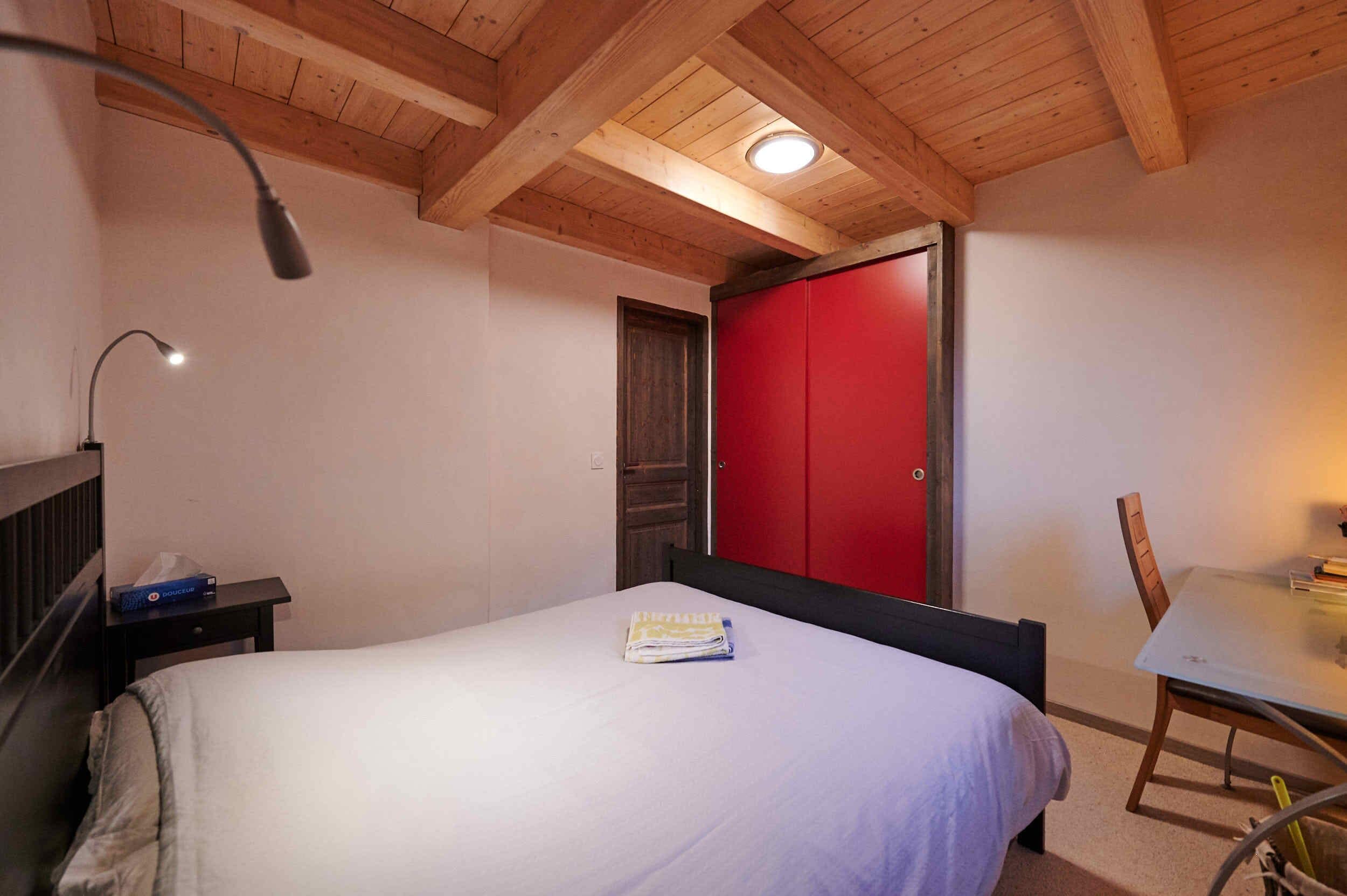 Ein Schlafzimmer mit einem Bett, einem Nachttisch, einem Schreibtisch mit Stuhl, einem roten Kleiderschrank und Holzbalkendecken.