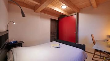 Een slaapkamer met een bed, een nachtkastje, een bureau met stoel, een rode garderobe en houten balken in het plafond.