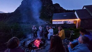 Un grupo de personas reunidas alrededor de una fogata en un entorno rústico al aire libre, con una montaña de fondo y un edificio que dice "Bed & Breakfast".