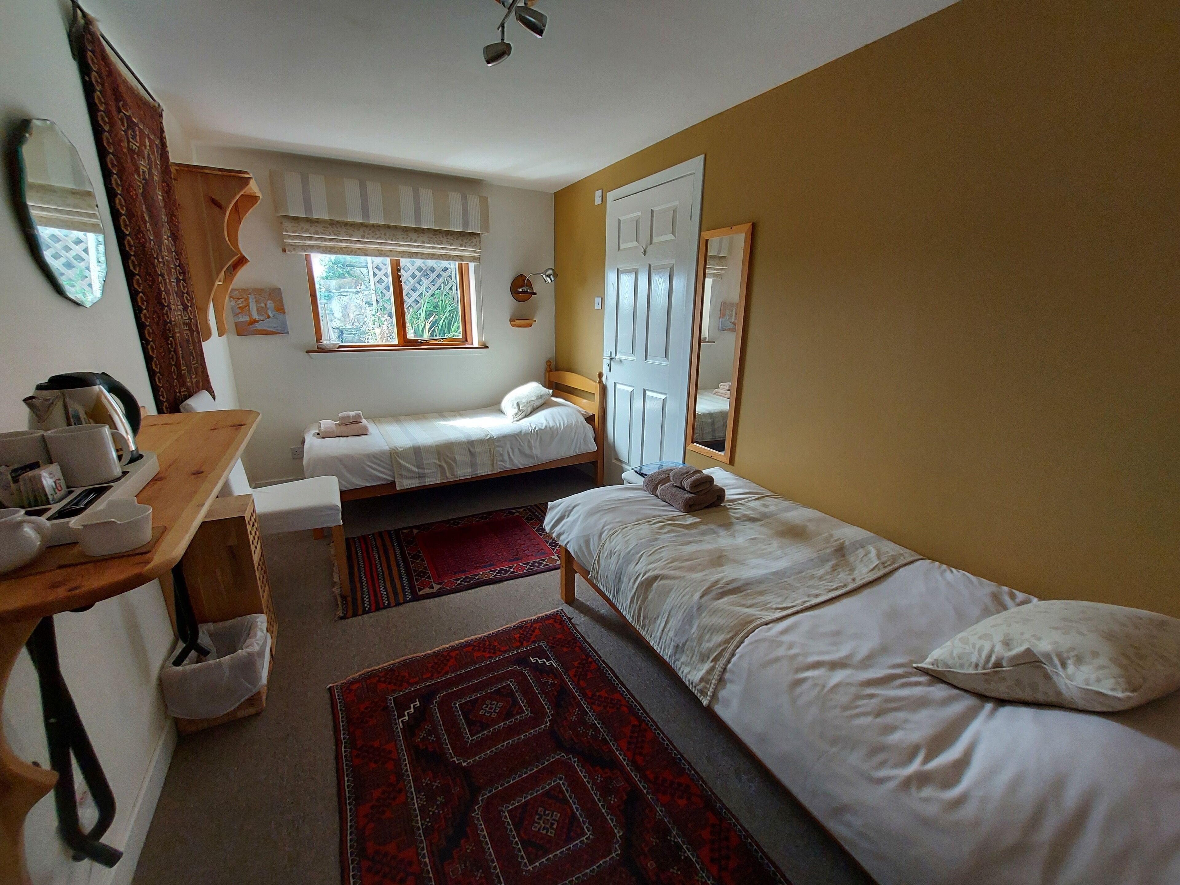 A small, cozy room with two single beds, a wooden desk with a kettle, a mirror, and a window with curtains.