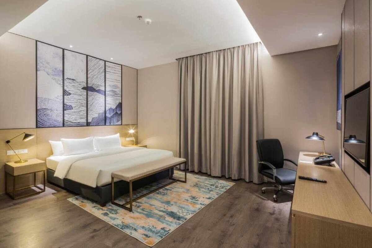 A modern hotel room with a large bed, bedside tables with lamps, a desk with a chair, a TV, and a large window with curtains.