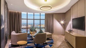 Une chambre d’hôtel moderne dotée d’une grande fenêtre donnant sur la ville, d’un luminaire rond au plafond, d’un téléviseur à écran plat et d’un coin salon agrémenté d’un tapis à motifs.