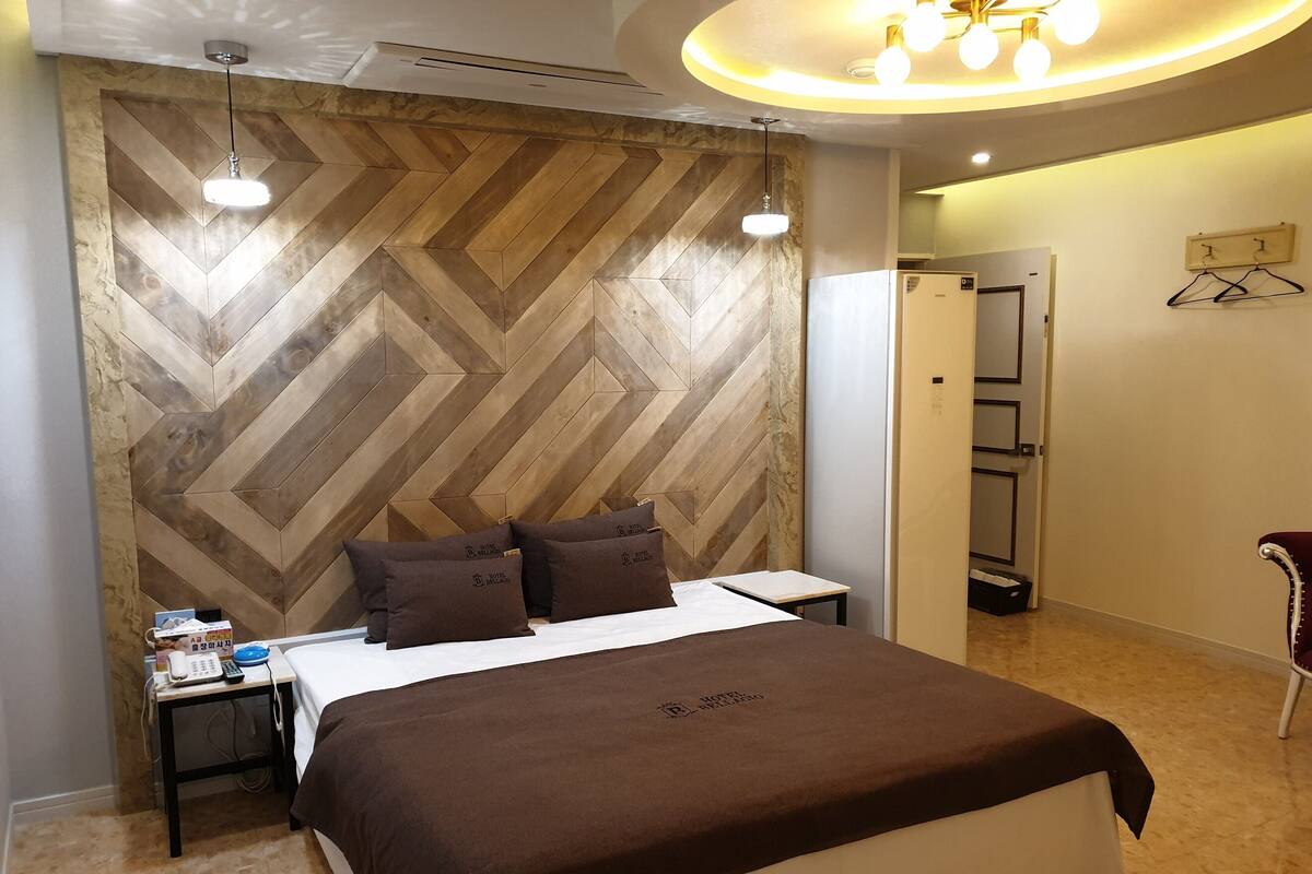 A hotel room with a herringbone-patterned headboard, a bed with a brown bedspread, bedside tables with a phone and remote control, and a hanging light fixture.