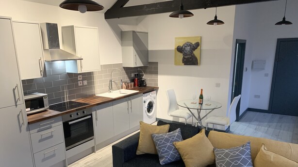 Une cuisine moderne avec des meubles gris, un four à micro-ondes et un lave-linge. On y trouve un canapé gris foncé avec des coussins à motifs, ainsi qu’une table en verre avec deux chaises. Un tableau représentant une vache est accroché au mur.