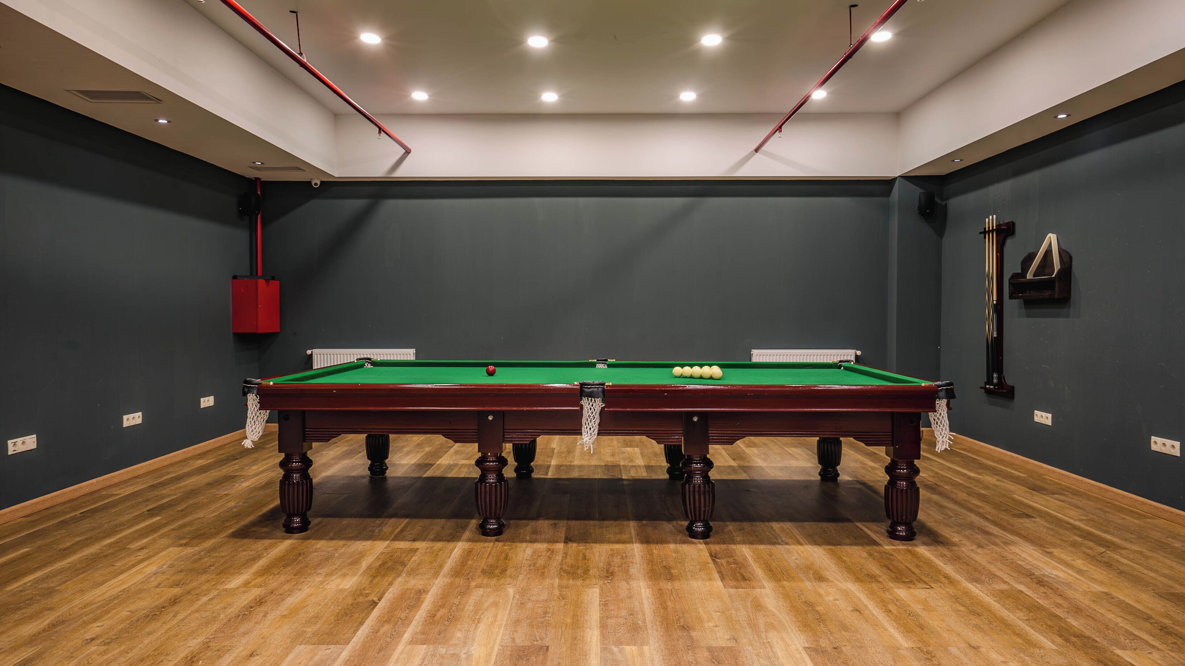 A pool table with balls, a red safety box, and a pool cue rack in a room with dark walls and wooden flooring.