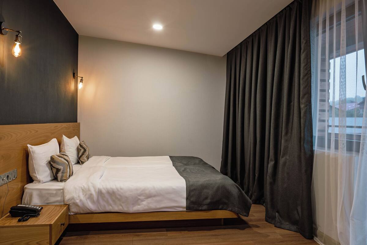 A modern hotel room with a wooden bed, a bedside telephone, and a wall-mounted light fixture.