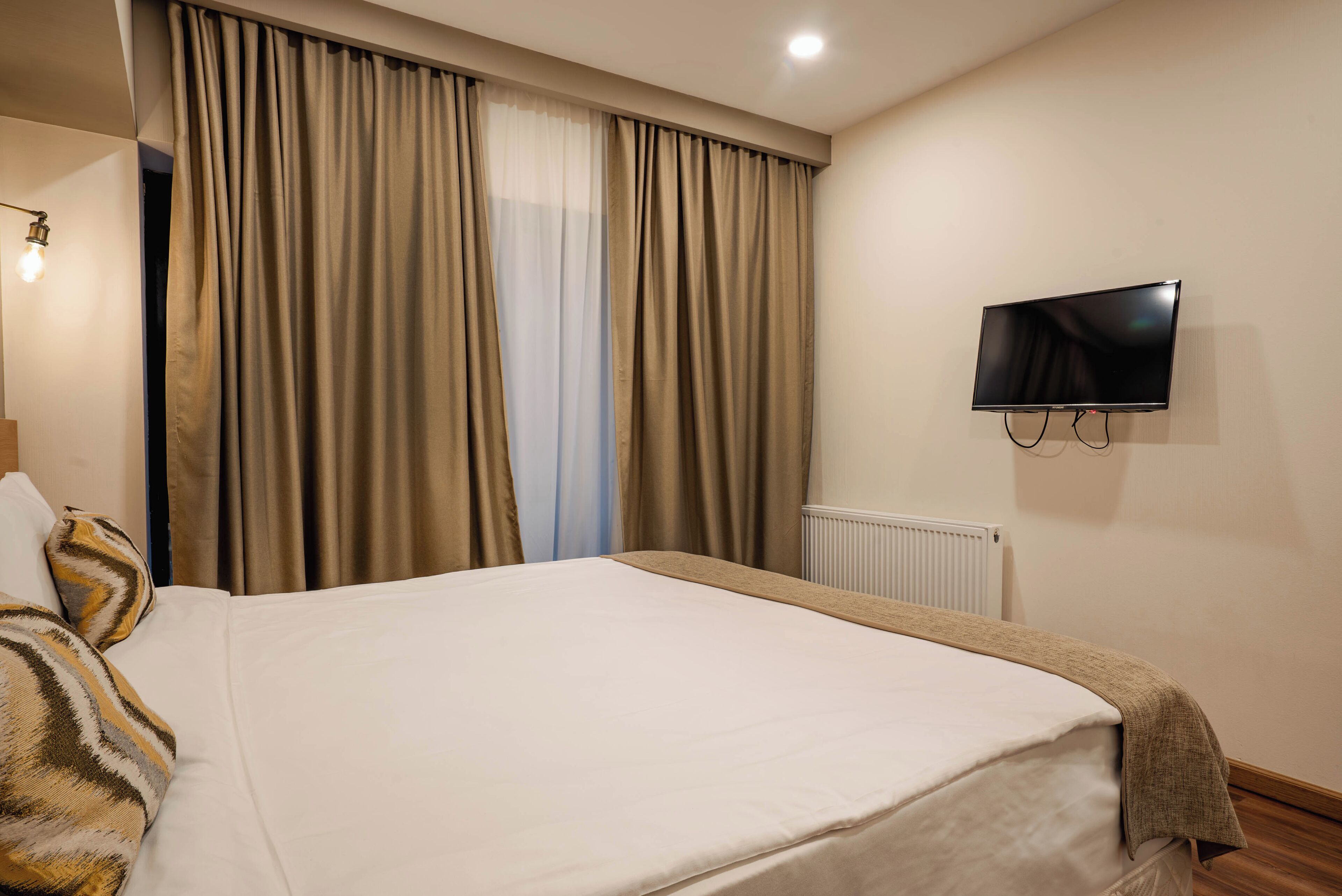 Une chambre d’hôtel avec un grand lit, des rideaux beiges, un téléviseur fixé au mur et un radiateur.
