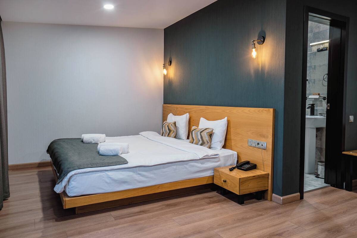 A modern hotel room with a wooden bed, white linens, a wooden nightstand, and a bathroom visible through an open door.
