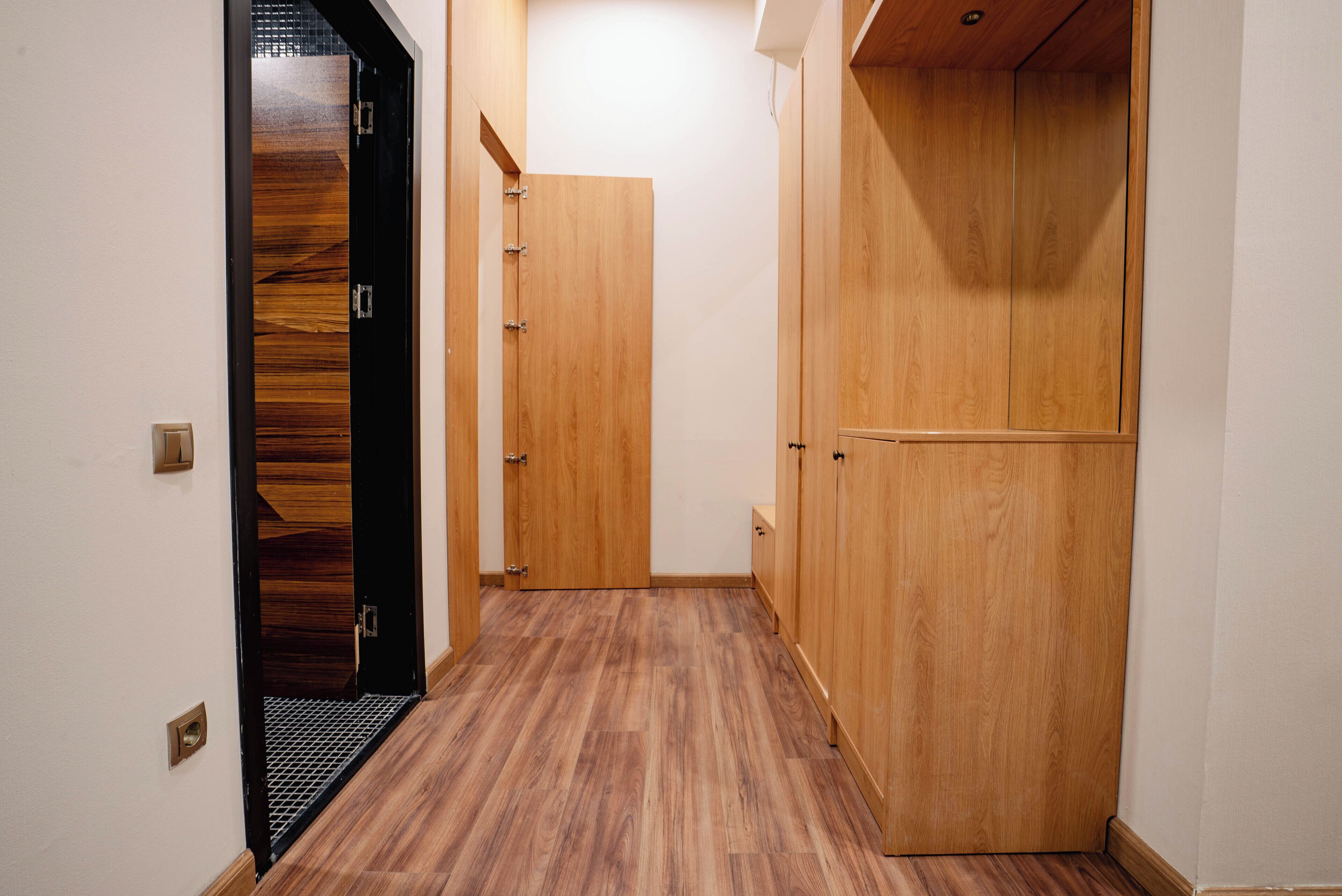 Un pasillo con piso de madera, una puerta negra con diseño texturizado y un armario de madera con pomo.