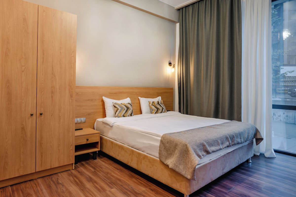 A hotel room with a wooden wardrobe, a bed with a headboard, two bedside tables, and a window with curtains.
