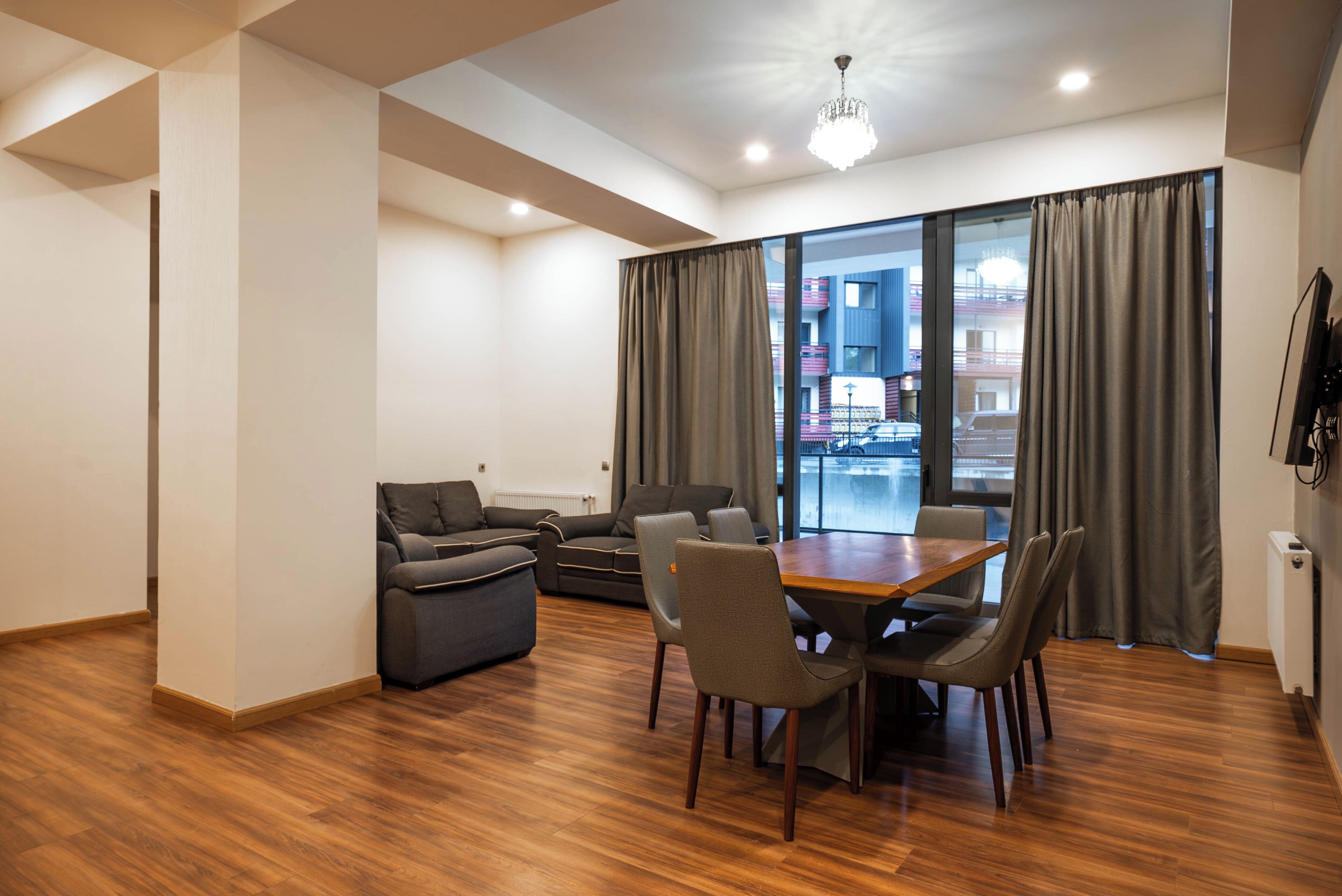 A modern living room with a dining area, large windows with curtains, and a chandelier.