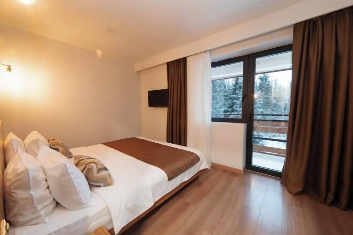 A bedroom with a bed, a window with curtains, a flat-screen TV, and a view of trees outside.