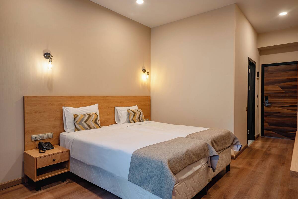 A hotel room with a wooden headboard, a bedside table with a phone, a bed with white bedding and a grey throw blanket, and two wall-mounted lights.