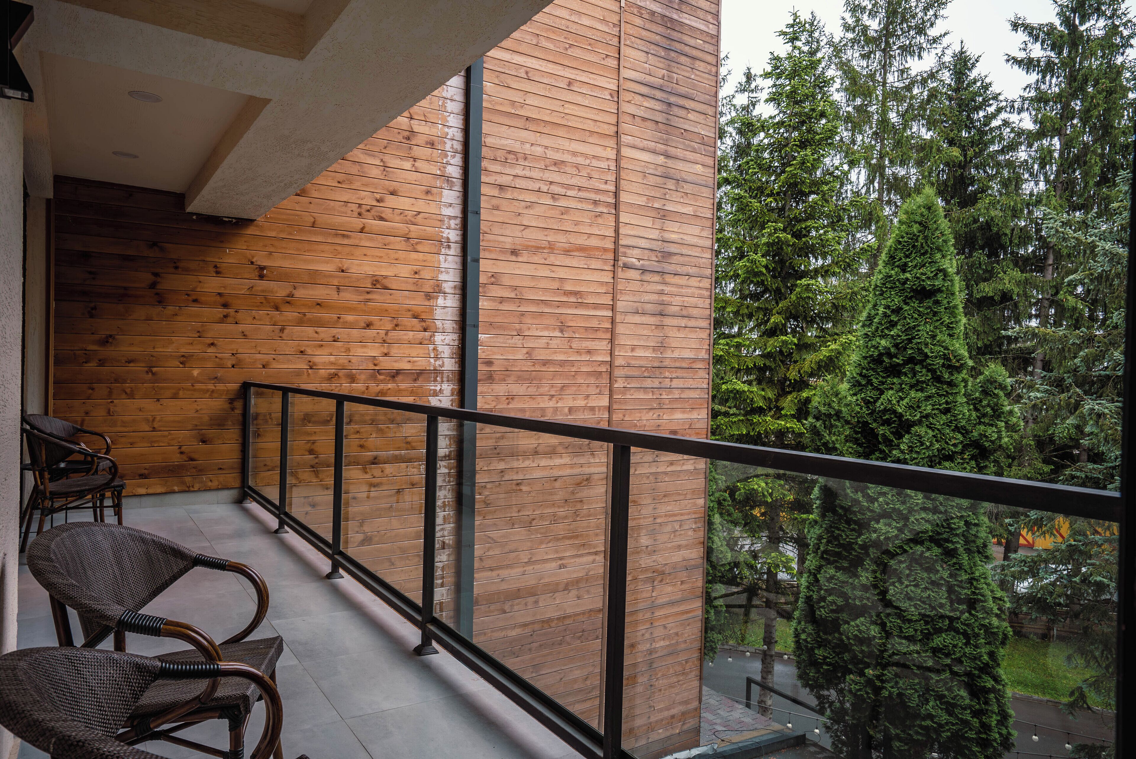 Un balcon en bois, agrémenté de chaises en osier, une rambarde en métal et une vue sur des arbres et une étendue d’eau.
