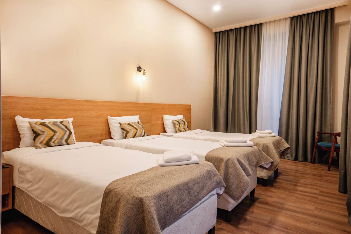 A hotel room with three beds, a wooden headboard, a wall-mounted light, and a view of a curtain.