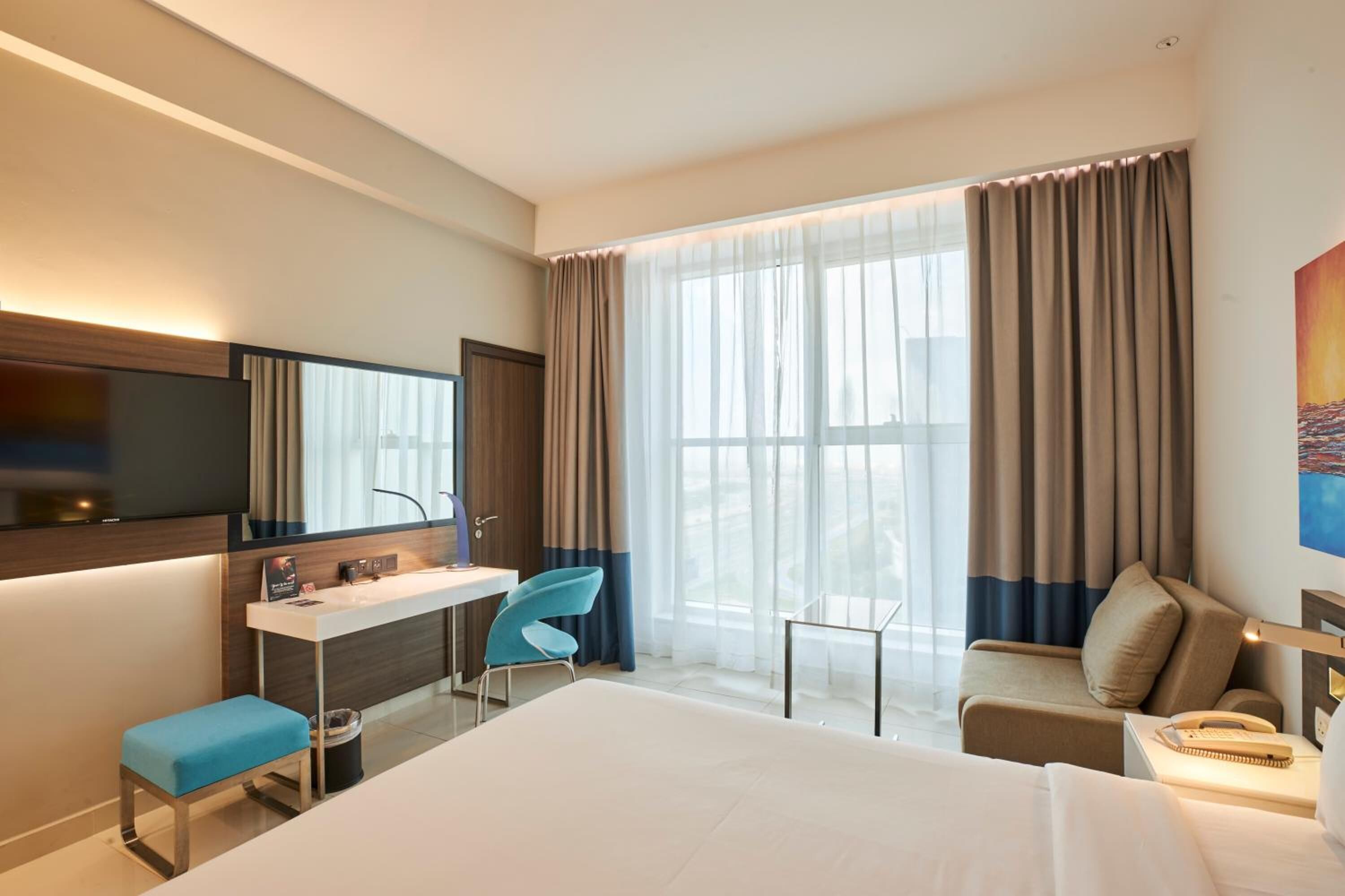 A modern hotel room with a large bed, a desk with a chair, a small table, a sofa, a TV, and a large window with curtains.
