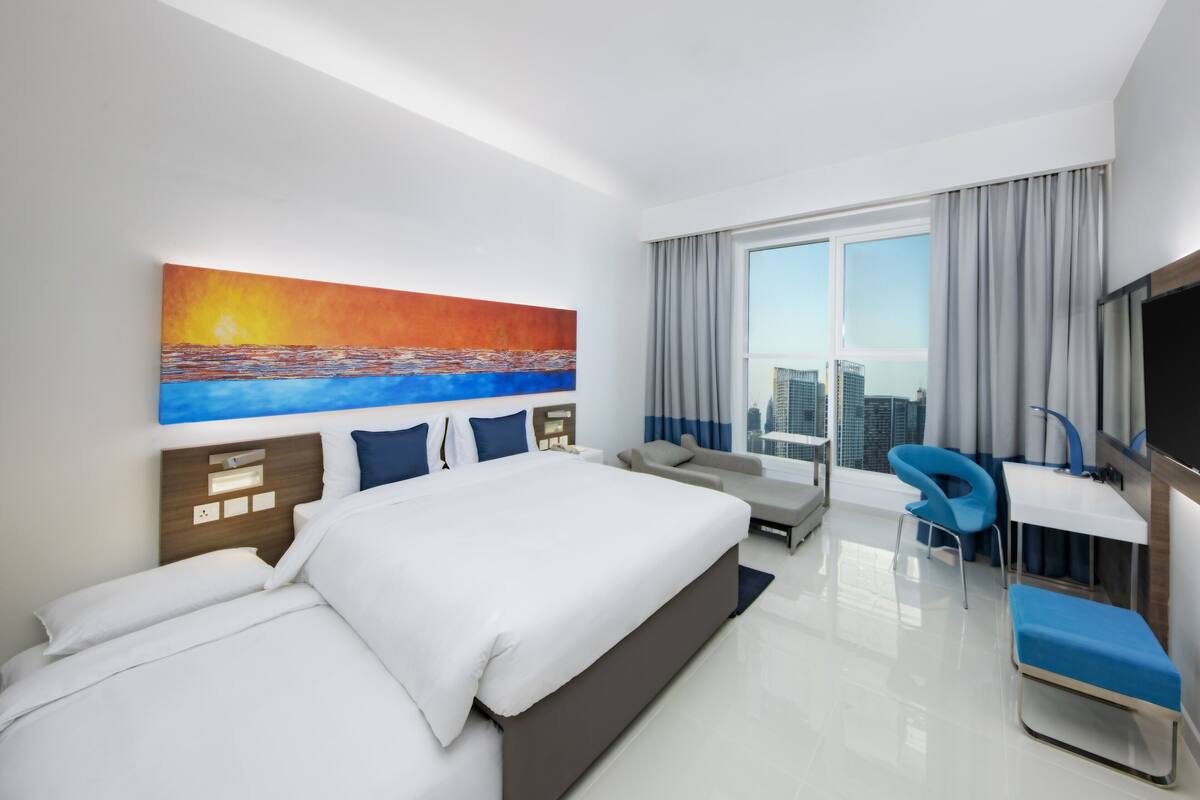 Une chambre d’hôtel moderne, dotée d’un grand lit, d’un bureau, d’une chaise et d’un tableau représentant un coucher de soleil sur l’océan.