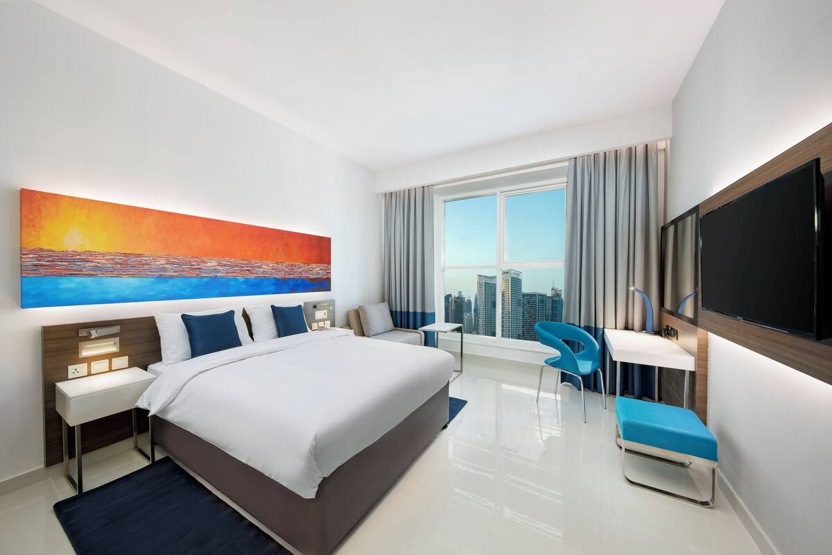 Une chambre d’hôtel moderne, dotée d’un grand lit, d’un bureau, d’une chaise, d’une télévision et d’un tableau représentant un coucher de soleil.