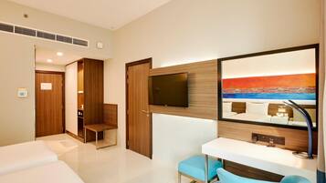 Quarto de hotel com cama, escrivaninha com cadeira, televisão na parede e um quadro com uma cena de praia.