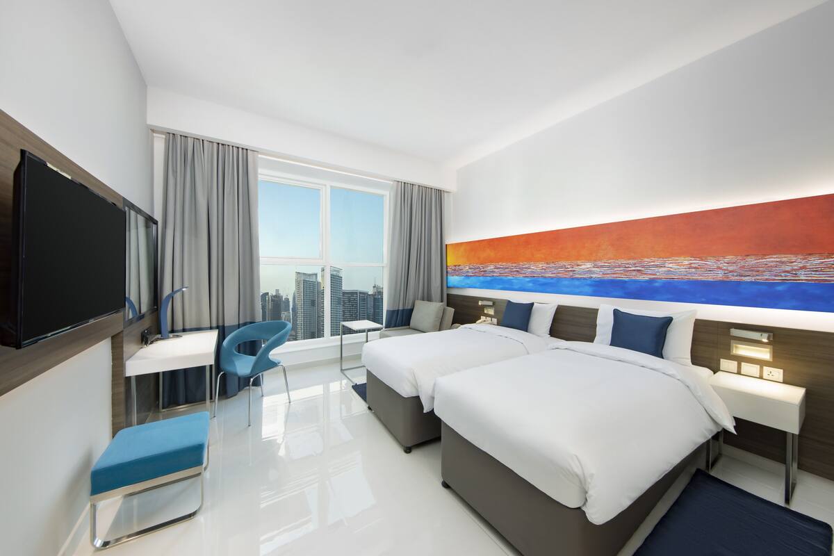 Une chambre d’hôtel moderne équipée de deux lits, d’un bureau, d’une chaise et d’une grande fenêtre donnant sur la ville.