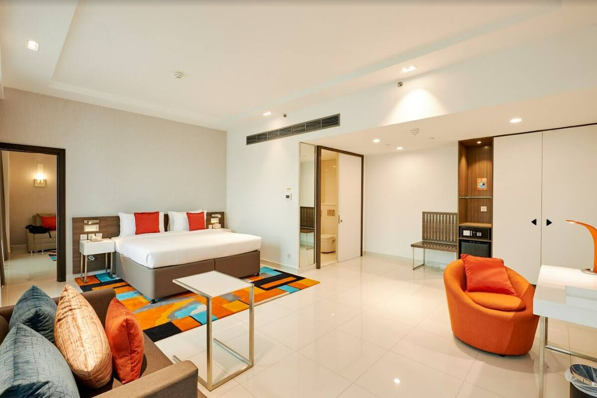 Une chambre d’hôtel moderne avec un grand lit, un canapé, un fauteuil orange et une petite table. On y trouve également un miroir, une table de chevet et une fenêtre.