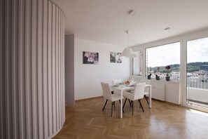 Un espace repas moderne avec une table et des chaises blanches, une grande fenêtre donnant sur la ville et un radiateur blanc.