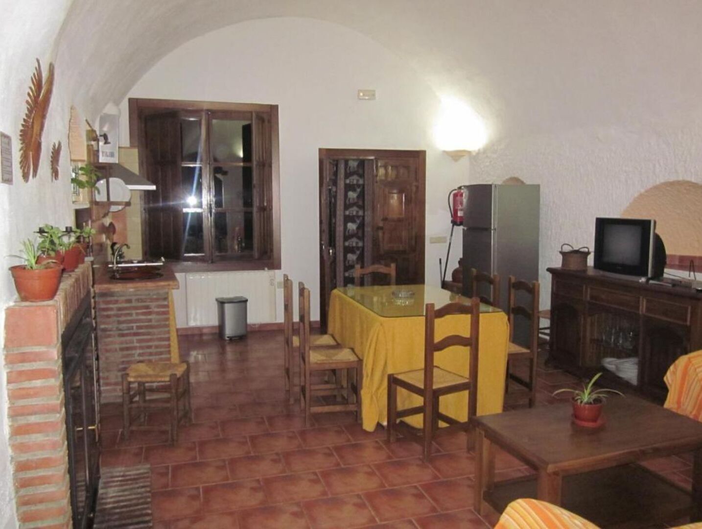 A dining area with a tiled floor, a table with yellow tablecloth, wooden chairs, a television on a cabinet, and potted plants.
