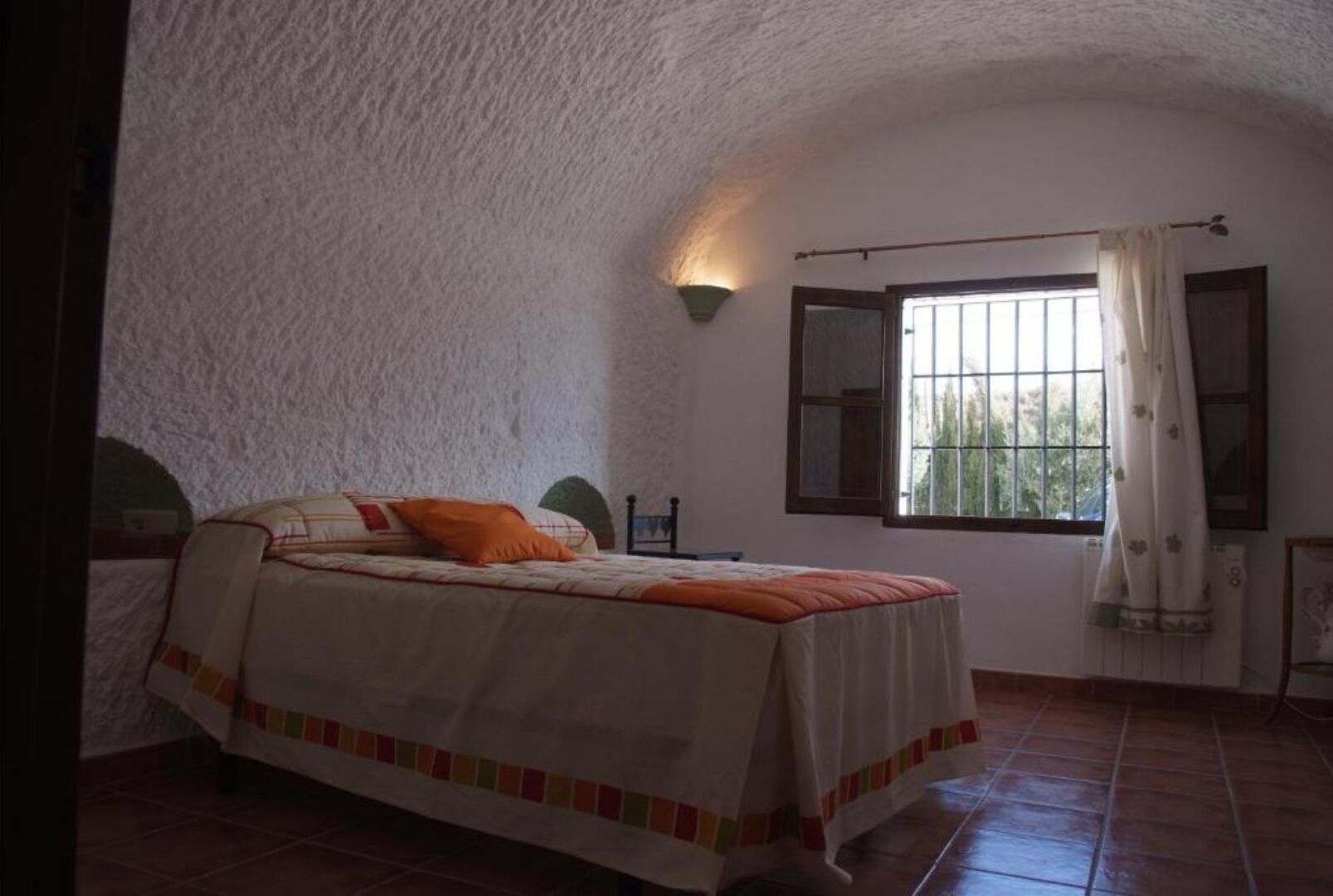 A bedroom with a bed, a window with curtains, a wall-mounted light, and a radiator.