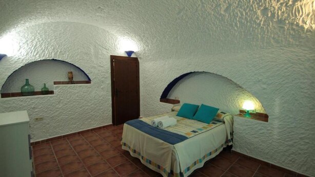 Ein Schlafzimmer mit gewölbter Decke, einem Bett mit blau-weißer Bettdecke, zwei Wandlampen und einem kleinen Regal mit Flaschen.
