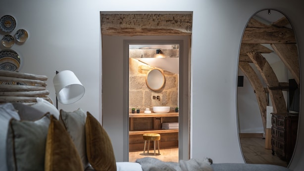 Une chambre à coucher comprenant un lit, une lampe de chevet, un miroir et donnant sur une salle de bain avec un lavabo et un tabouret.