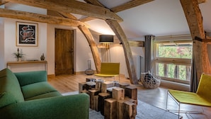 Un intérieur rustique avec des poutres apparentes, un canapé vert, un fauteuil jaune et une grande fenêtre donnant sur la forêt.
