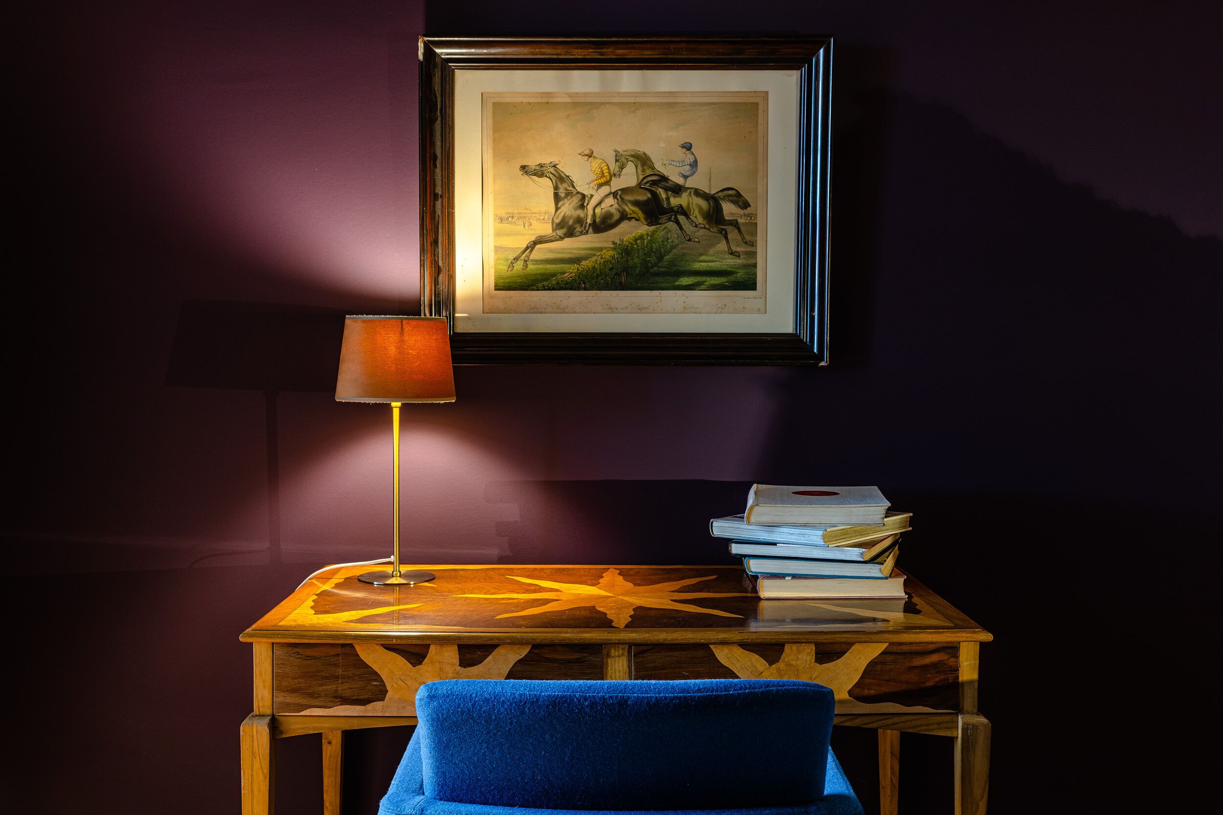 Een lijst met een afbeelding van paarden aan een muur, een tafel met een stapel boeken en een blauwe stoel.