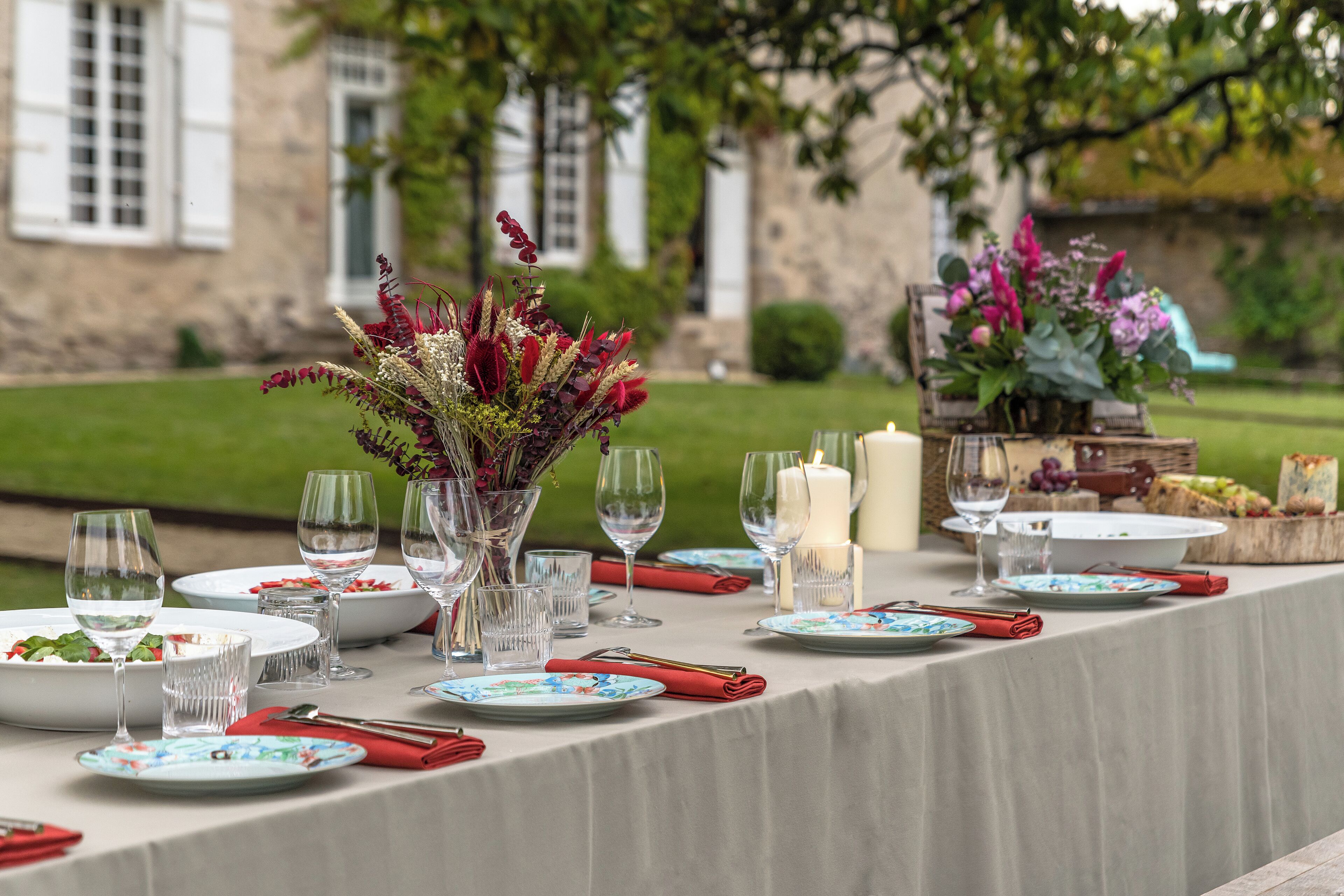 A table set for a meal with plates, glasses, and cutlery, surrounded by decorative flowers and candles, set against a stone building backdrop.