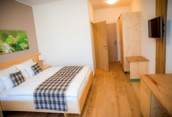 A bedroom with a wooden bed, a bedside table, a TV mounted on the wall, and a door leading to another room. - Gasthof Wachter (Gaal)