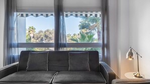 Ett modernt vardagsrum med en grå soffa och fåtölj, ett litet sidobord och en golvlampa. Ett stort fönster med tunna gardiner ger utsikt över palmer och grönskande växter.