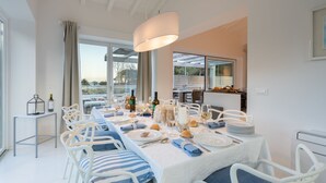 Uma sala de jantar com uma mesa posta para quatro pessoas, cadeiras brancas, um lustre e vista para o exterior.