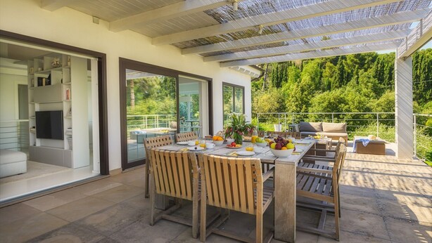 Un espace de restauration extérieur moderne, avec une table dressée pour le petit-déjeuner, entouré de chaises en bois et d’une terrasse fermée par des parois de verre.