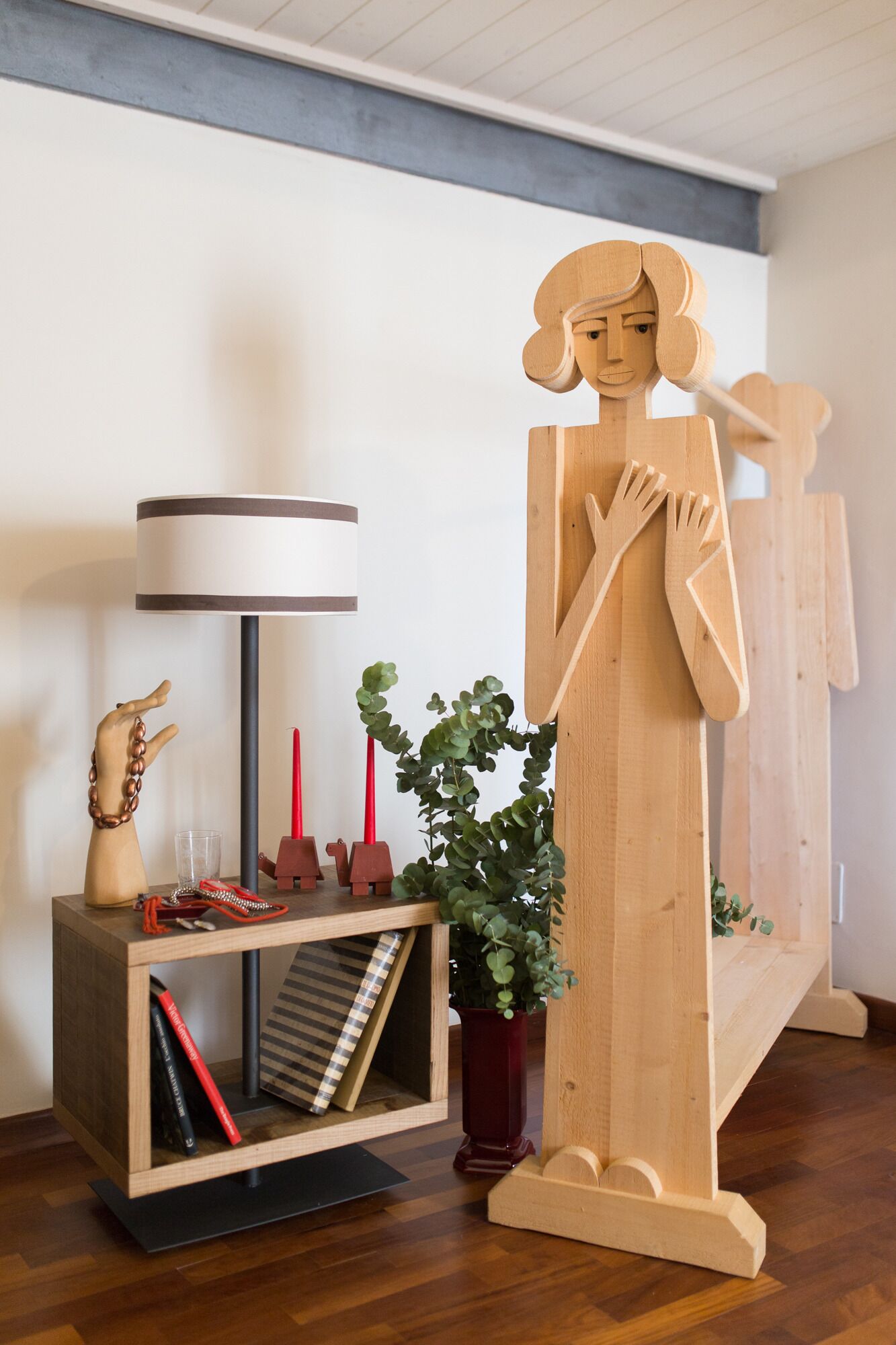 A wooden sculpture of a figure with a long robe, a lamp, a plant, and books on a wooden shelf.