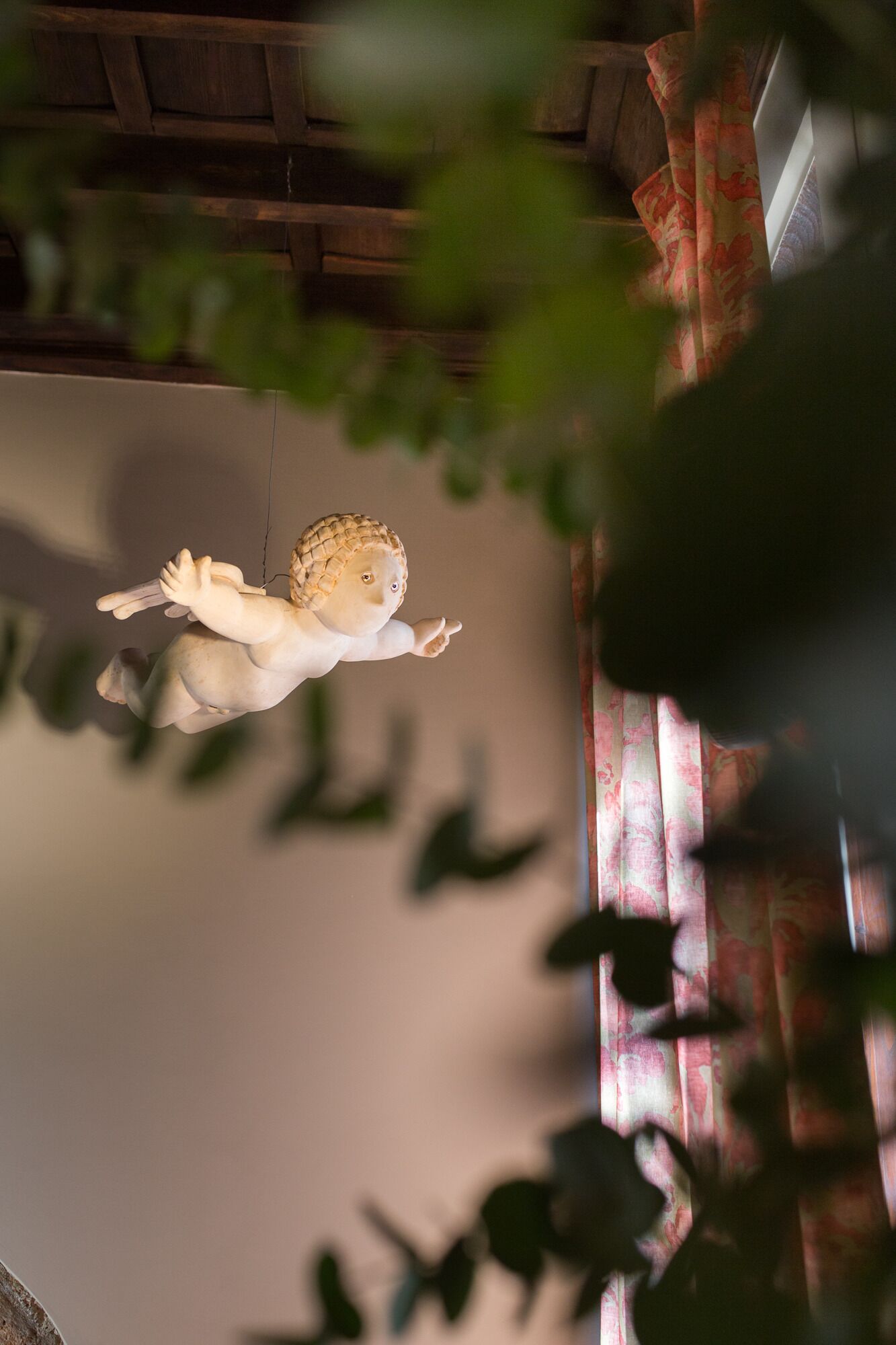 A statue of a figure with outstretched arms, positioned indoors with a wooden ceiling and a patterned curtain in the background.
