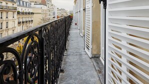 Un balcón con barandilla de hierro forjado, un edificio con contraventanas blancas y vista a otros edificios al fondo.
