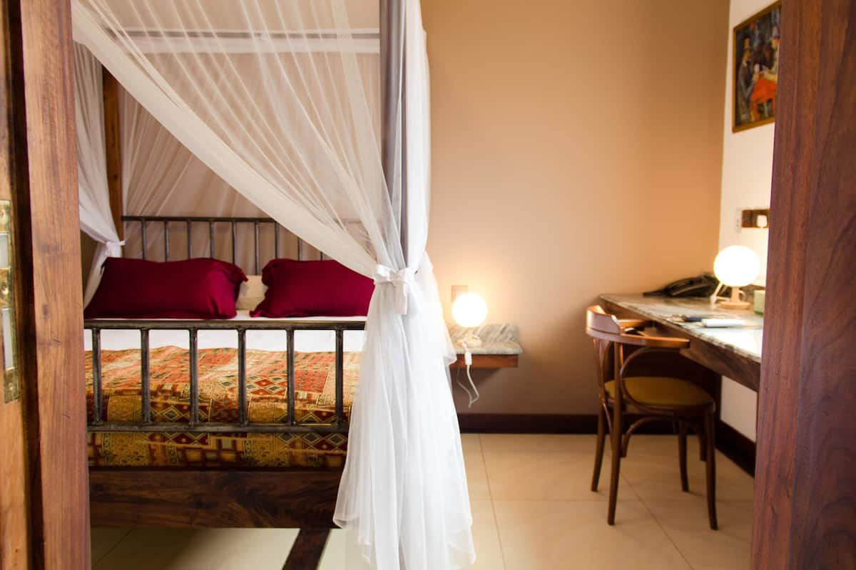 Ein Zimmer mit einem Bett unter einem Moskitonetz, einem Schreibtisch mit Stuhl und einem an der Wand befestigten Regal.