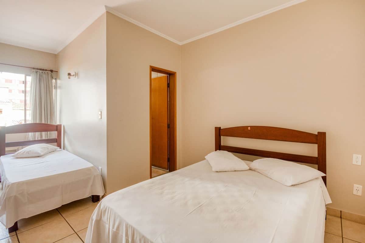 A hotel room with two single beds, a wooden headboard, a window with curtains, and a door leading to another room.