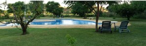 A swimming pool with a circular shape, surrounded by grass and trees. There are two chairs and a table nearby.