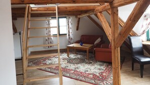 Een loftachtige kamer met zichtbare houten balken, een houten ladder, een rode bank, een klein tafeltje en een kamerbreed tapijt met een patroon.