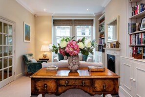 Ein Zimmer mit Schreibtisch aus Holz, Blumen in einer Vase, einem Bücherregal und einem Kamin.
