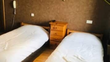 Twee enkele bedden met houten frames en witte beddengoed, een nachtkastje van hout, en een stopcontact aan de muur.