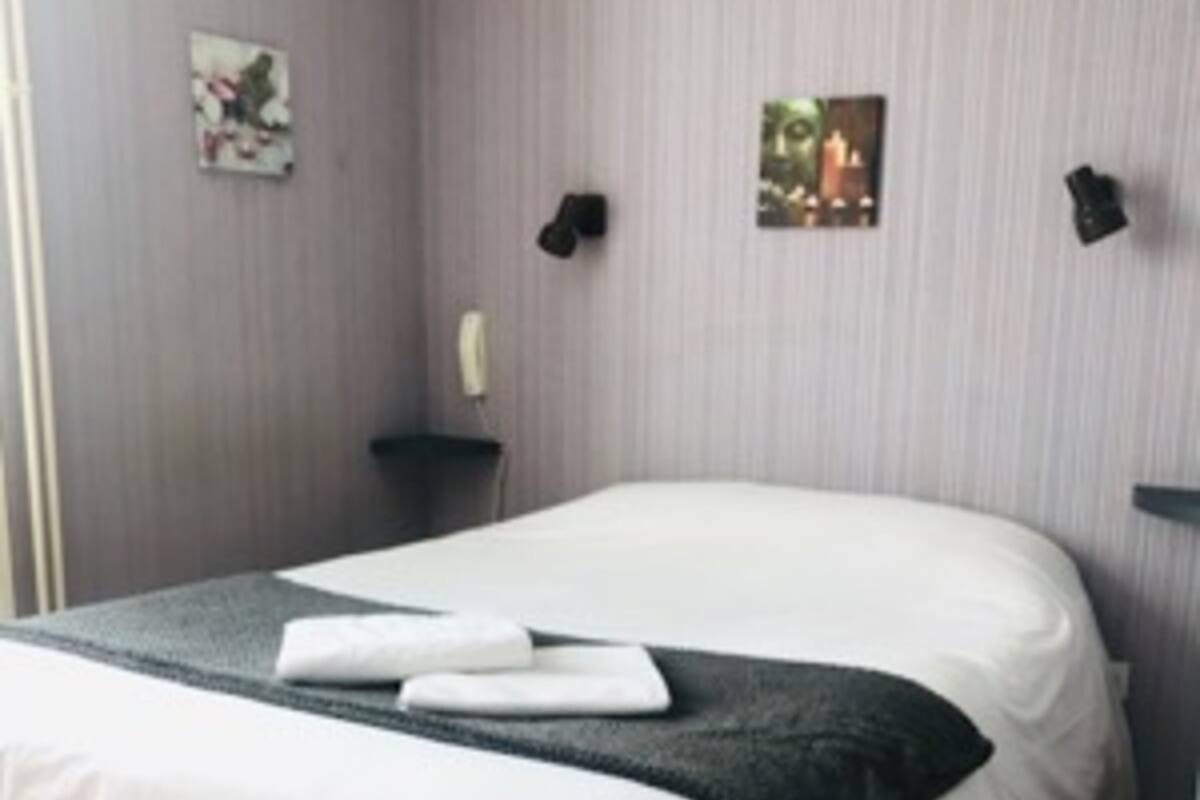 A neatly made bed with white linens and a dark gray blanket, a wooden floor, and a wall with two framed pictures and two wall-mounted lamps.